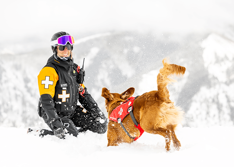 Aspen Snowmass Patroller kneels on the ground as her golden retriever patrol dog bounds towards her in the snow