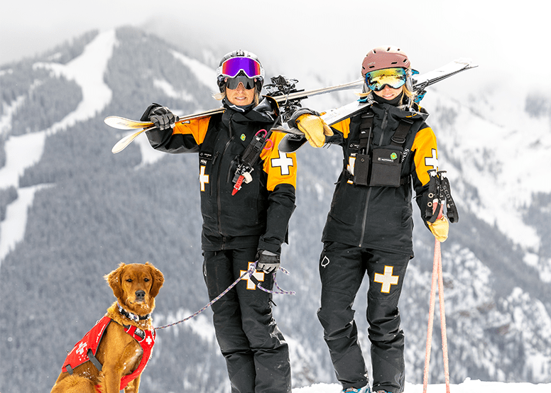 Two female patrollers stand on Buttermilk with their skies on their shoulders and a golden retriever patrol dog sits patiently next to them