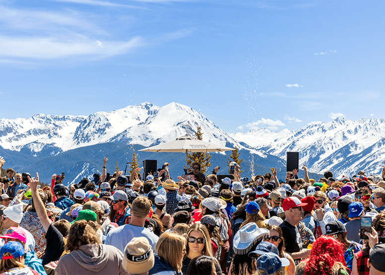 crowd gathers under blue skies, with huge snowy mountains in the background, as a DJ plays atop aspen mountain