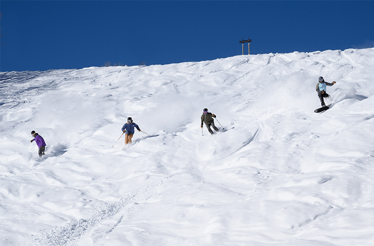 Four skiers and snowboarders ride down a hill in deep powder on a blue bird day at aspen Snowmass