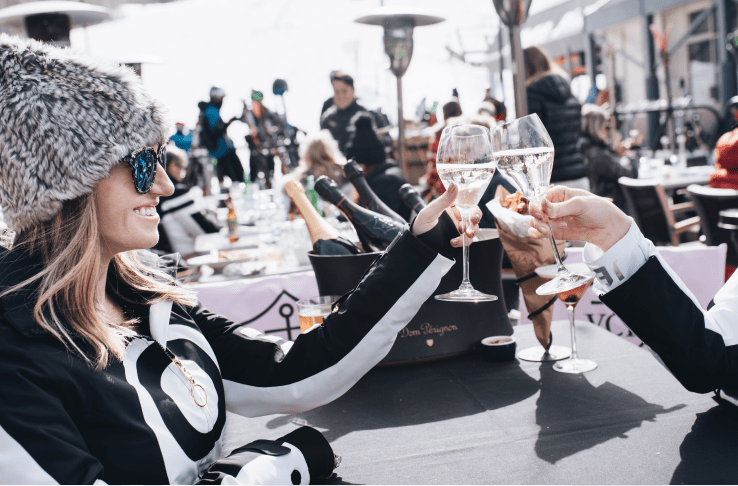 two women in black and white knit sweaters cheers their wine glasses at the base of Aspen sKI rESORT, AT aJAX tAVERN