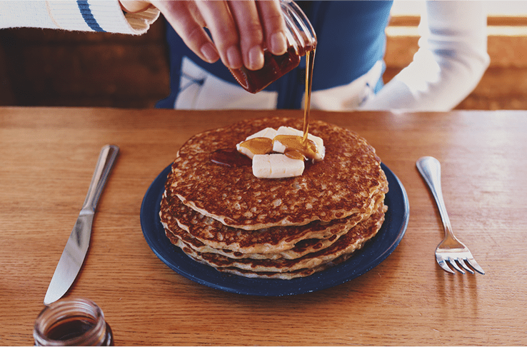 Stack of pancakes on wood table, slabs of butter on top an woman pours syrup over the top