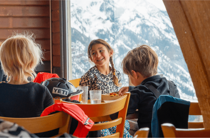 Three kids laugh at a table at the Buttermilk Cliff house on a snowy day, their ski gear fills the table