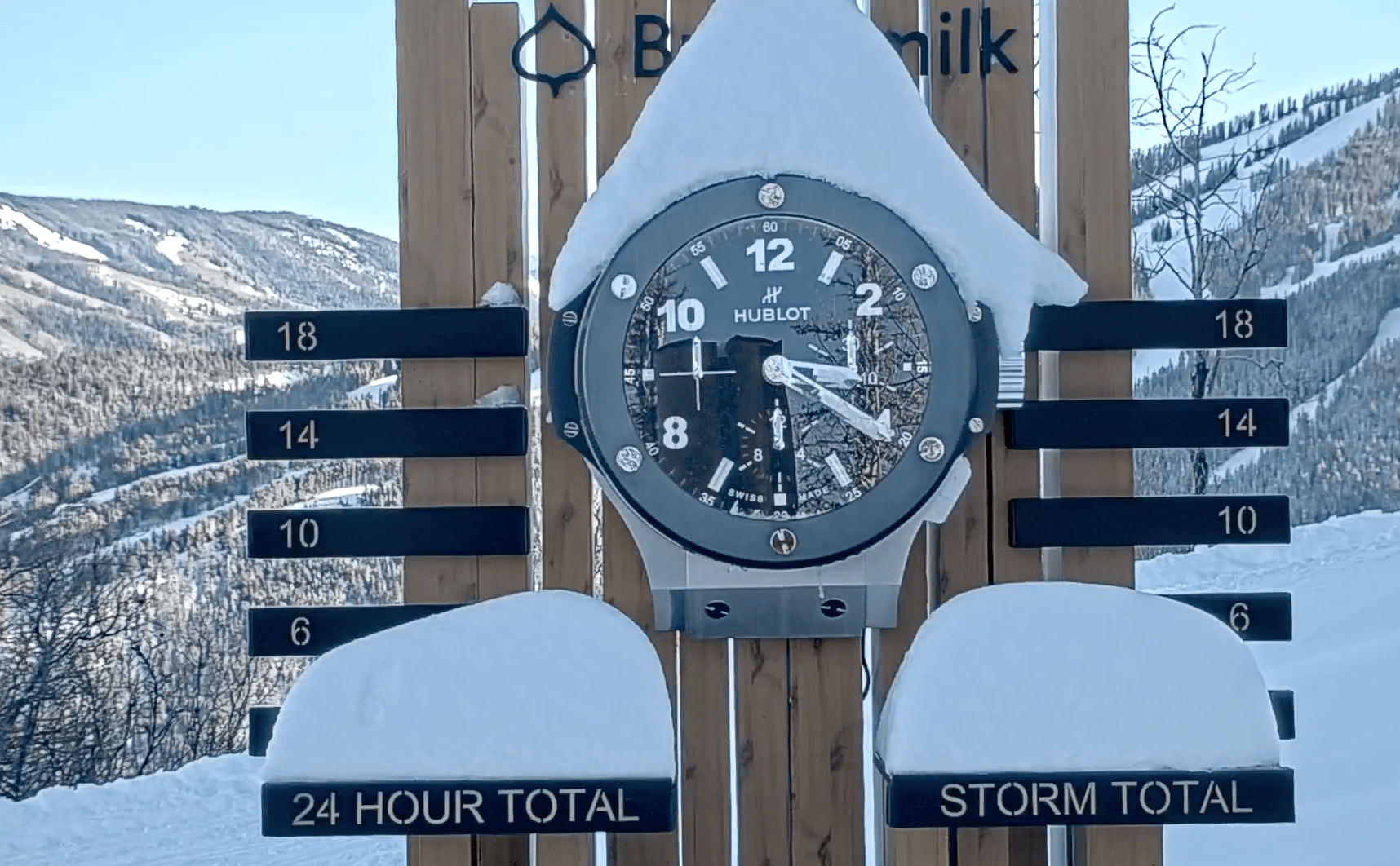 Buttermilk snow stake, in the shadow of the mountain with blue skies behind, snow piled on the top of the clock and up to the 6 inch mark on the stake