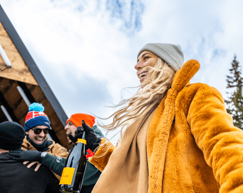 Woman in a burnt orange fuzzy jacket and beanie dances with a bottle of champagne in her hand