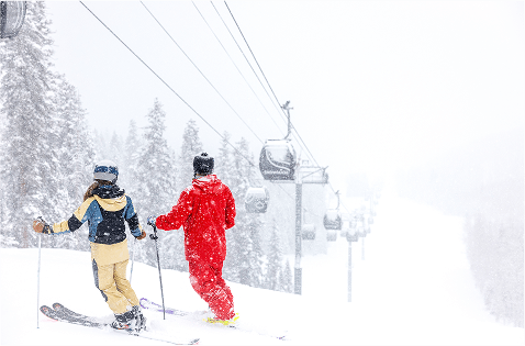a ski pro and their guest stand on the snowy slope, looking down at the snowmass gondola