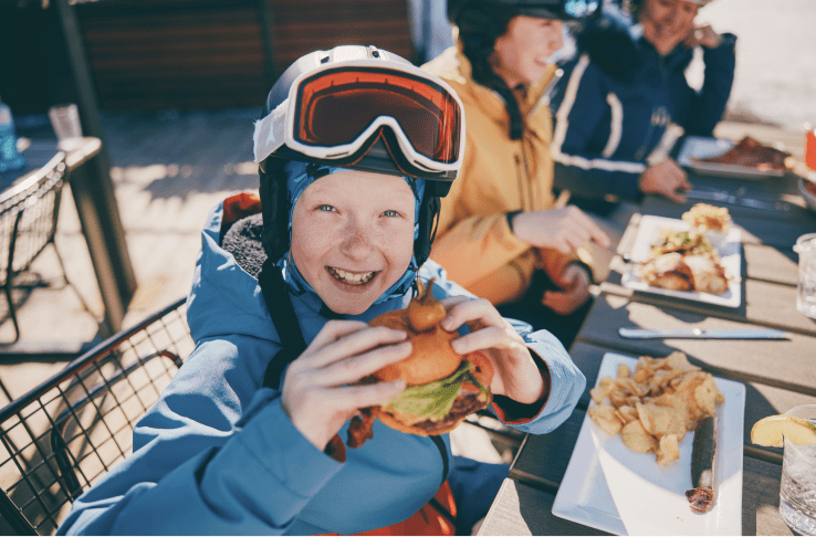 Little boy smiles at the camera in his ski gear as he holds a big cheese burger at lunch at aspen snowmass