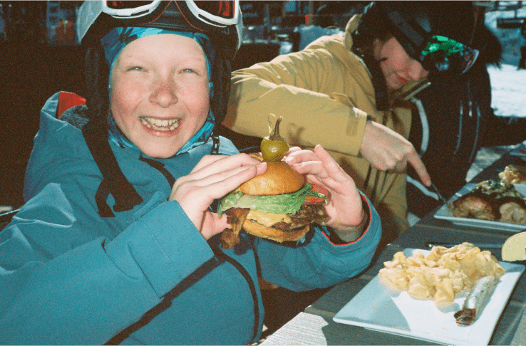 Little boy smiles as he holds a large burger, ready to take a bite, in his ski gear at Aspen Snowmass
