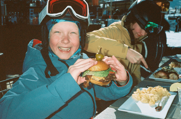 Little boy smiles as he holds a burger in his ski gear