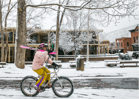 man bikes through snowy town with his skies on his shoulder