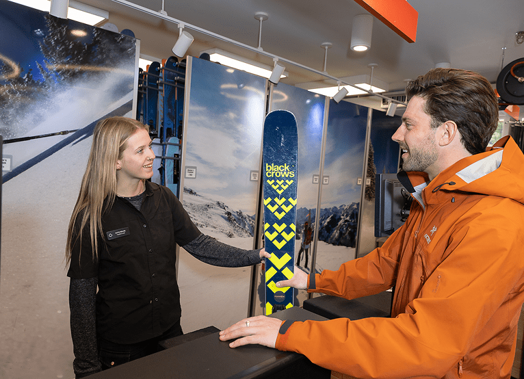 Woman working at Snowmass Four Mountain Sports smiles at customer as she hands him his ski rentals for the day