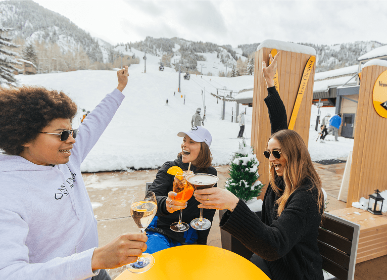 Three people cheers their mixed drinks on a cloudy day at the base of Aspen Mountain, at ajaz tavern