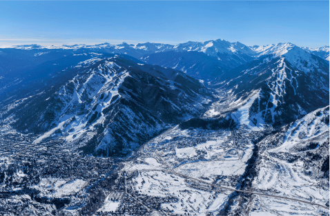 Areal view of Aspen, ski mountain towers over the town and blue sky illuminates the expansive mountain range behind aspen mountain