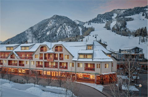 the little nell glows with warm light through each homey window on a cold winter night, the aspen mountain ski resort can be seen with pink skies behind