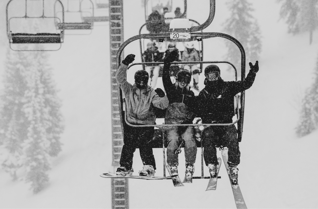 Black and white image of three people on a chairlift, arms raised as they cheer for the camera at Aspen Snowmass