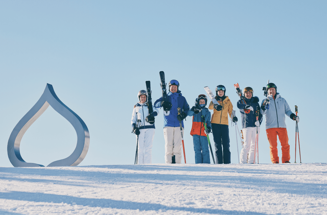 Family of 6 stands with their skies on their shoulder next to an Aspen Snowmass leaf installment