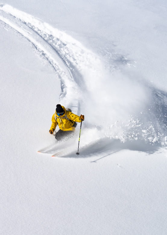 A skier in a bright yellow jacket carves through fresh, powdery snow, creating a trail of spray behind them.