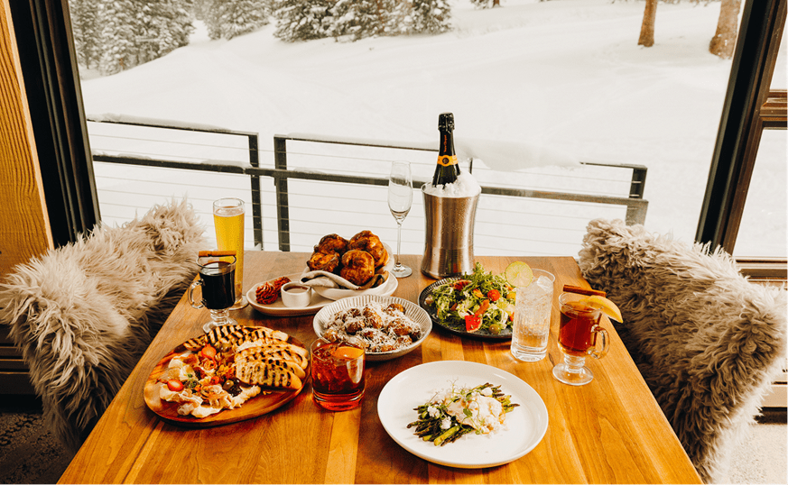 Wood table with plates of delicious food, appetizers, main courses, and craft cocktails at the Alpin Room Snowmass
